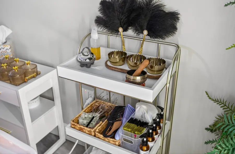 A white trolley of head-spa tools — brass singing bowls, gua sha stones, oils and a black feather fan