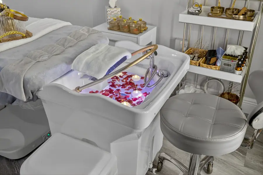 A white head-spa basin scattered with rose petals, a brass bowl of tools and a stylist stool alongside