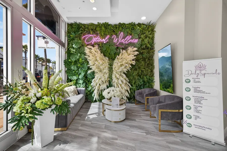 Pink neon Optimal Wellness script mounted on a green living wall between two tall white pampas and floral wing installations