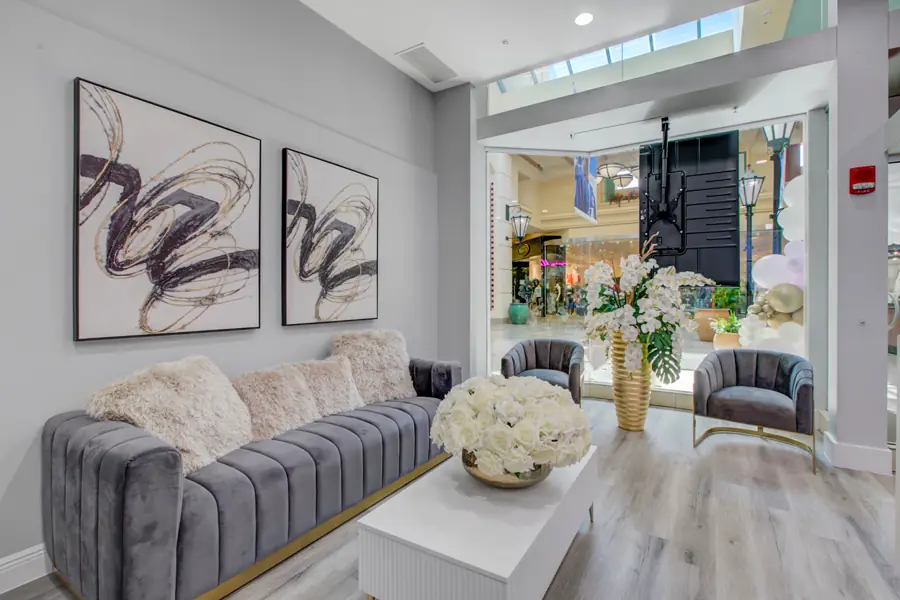 A grey velvet sofa and gold-framed armchairs with white hydrangeas, looking out through glass to the walkway