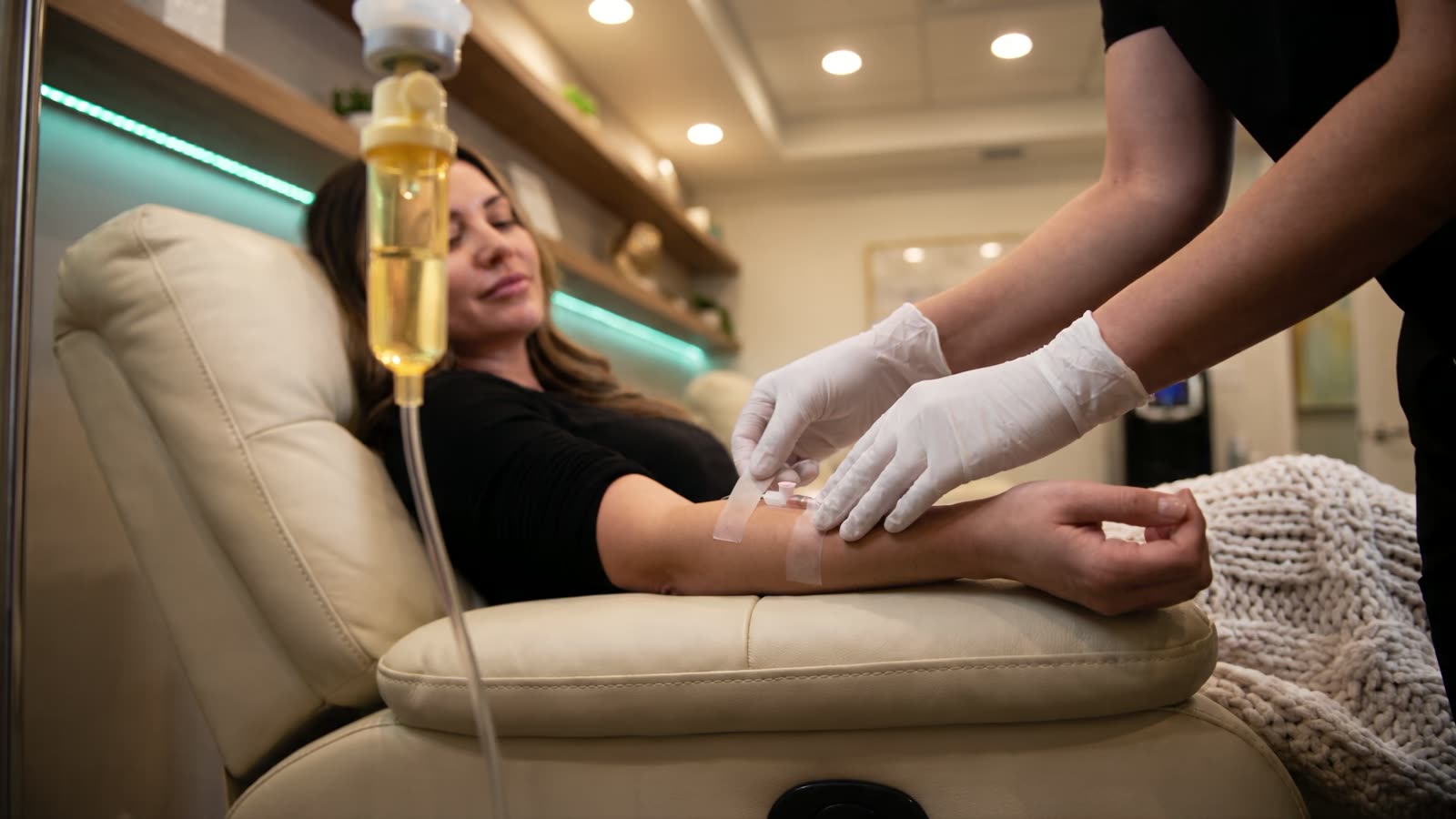 A nurse in white gloves smoothing tape over an IV line on a relaxed client's forearm in a plush lounge chair